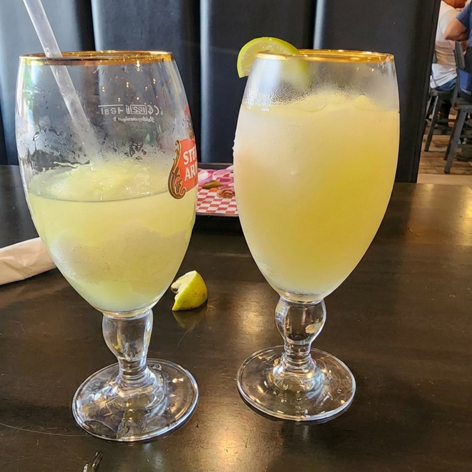 Two frosty pale-yellow citrus drinks in stemmed glasses sit on a restaurant table, garnished with lime wedges.